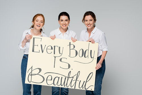 Cheerful women holding placard with every body is beautiful lettering isolated on grey