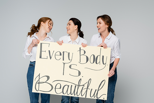 Smiling women looking at each other while holding placard with every body is beautiful lettering isolated on grey