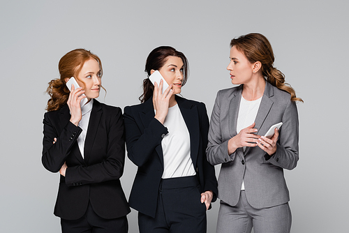 Businesswomen looking at colleague while talking on smartphones isolated on grey