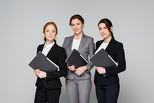 Positive businesswomen holding paper folders isolated on grey