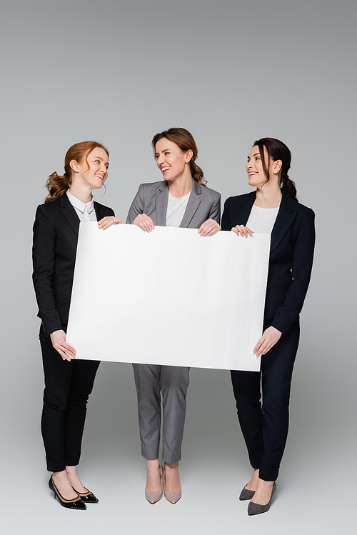 Businesswomen with empty white board smiling at each other on grey background