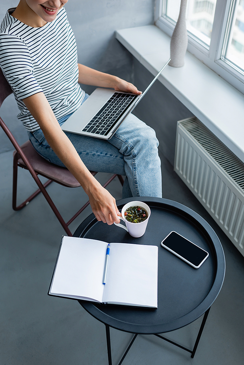 Cropped view of freelancer holding cup of coffee while using laptop near smartphone and notebook at home