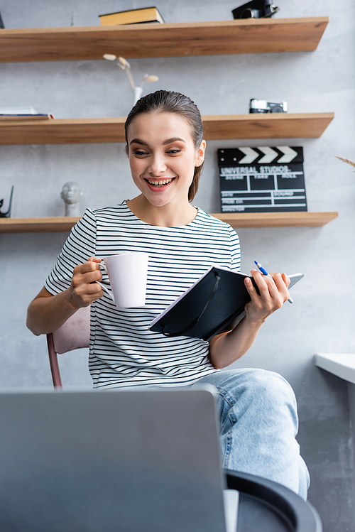 Selective focus of brunette teleworker with cup and notebook looking at laptop while working from home
