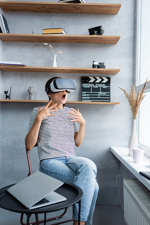 Selective focus of shocked woman in vr headset sitting near laptop on coffee table in living room