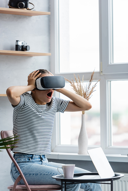 Selective focus of excited woman using vr headset near cup and notebook on coffee table at home