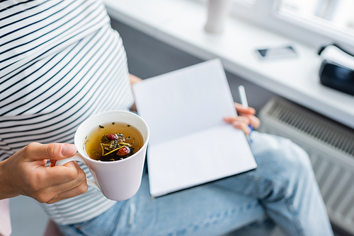 Cropped view of woman holding cup of herbal tea and notebook at home