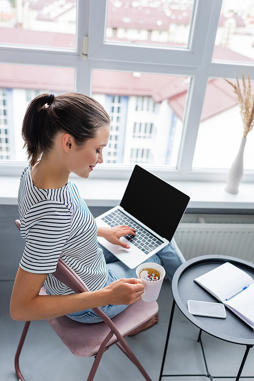 Brunette freelancer using laptop while holding cup of herbal tea near notebook and smartphone on table