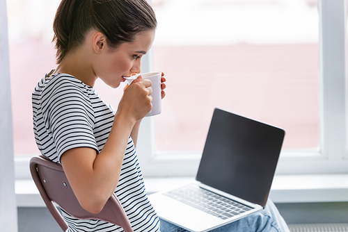 Selective focus of woman drinking tea and looking at laptop with blank screen at home