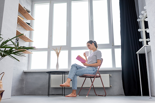 Selective focus of barefoot woman with cup reading book while sitting near window at home