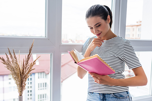 Young woman reading book near window at home