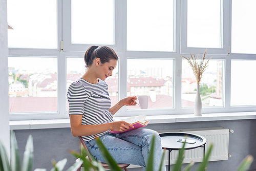 Selective focus of woman with cup reading book near smartphone and coffee table at home