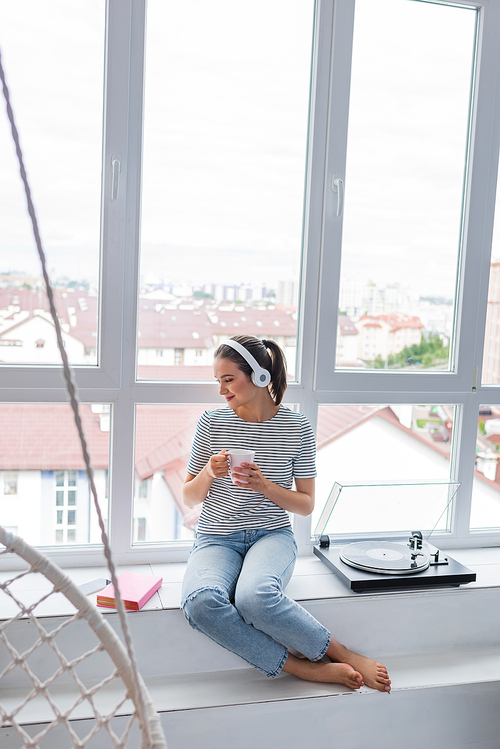 Selective focus of young woman in headphones holding cup near book and vinyl player on windowsill