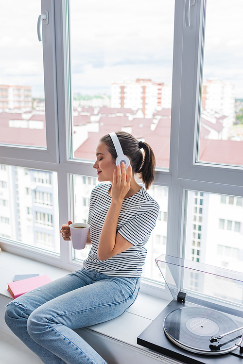 Selective focus of woman listening music in headphones while holding cup near vinyl player on windowsill
