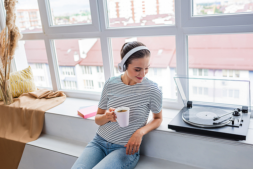 Woman with cup listening music in headphones near vinyl player on windowsill