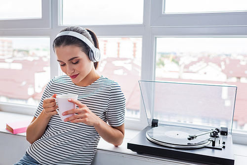 Young woman in headphones holding cup near vinyl player on windowsill