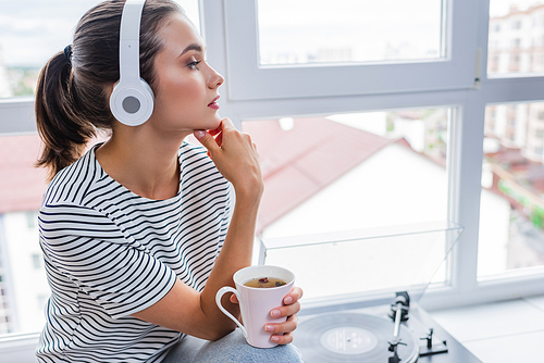 Young woman with cup of tea listening music in headphones near vinyl player on windowsill