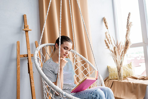 Young woman reading book while sitting in hanging armchair at home