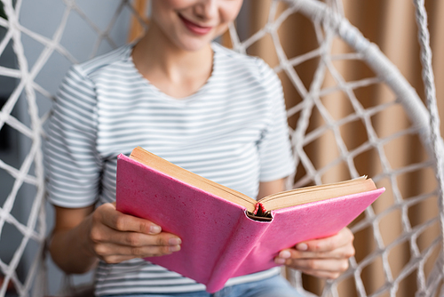 Cropped view of young woman reading book in hanging armchair at home