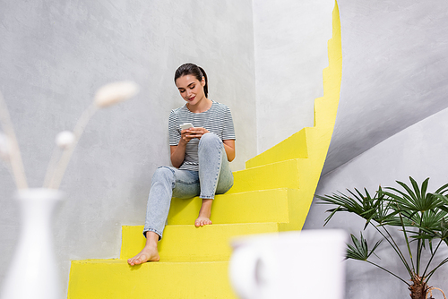 Selective focus of barefoot woman using smartphone on stairs at home