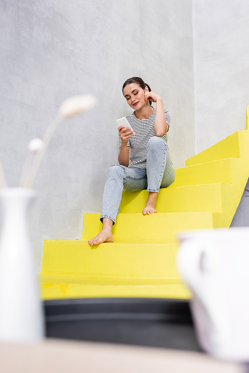 Selective focus of barefoot woman using smartphone while sitting on stairs at home
