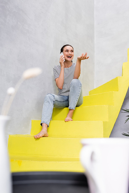 Selective focus of excited woman talking on smartphone while sitting on ladder at home