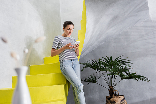 Selective focus of shocked woman using smartphone near stairs at home