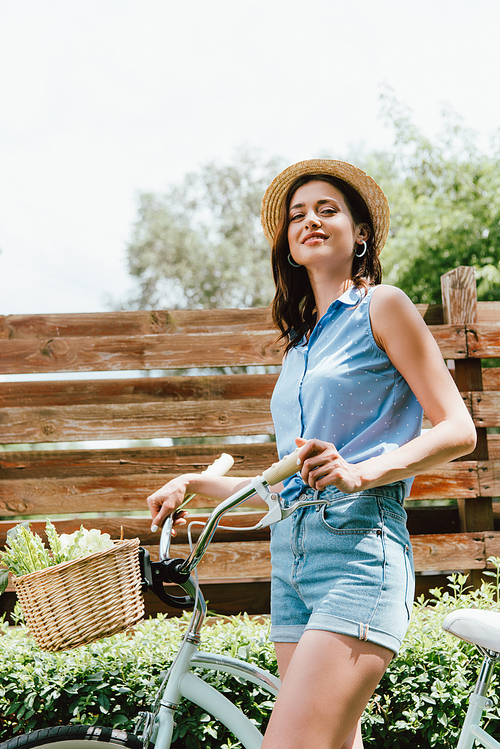 cheerful girl in straw hat standing with bicycle outside