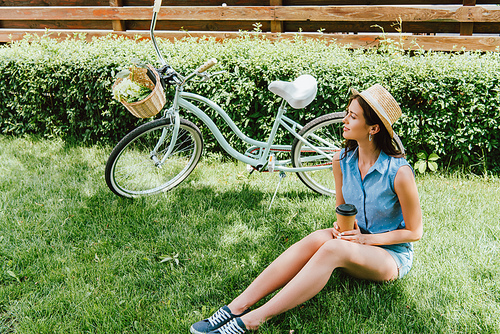 woman in straw hat holding paper cup and sitting on grass near bicycle