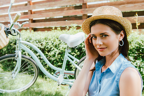 attractive woman touching straw hat and smiling near bicycle