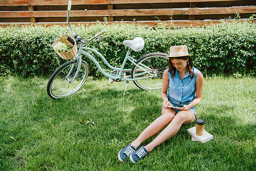 girl in straw hat using digital tablet and sitting on grass near bicycle, paper cup and books