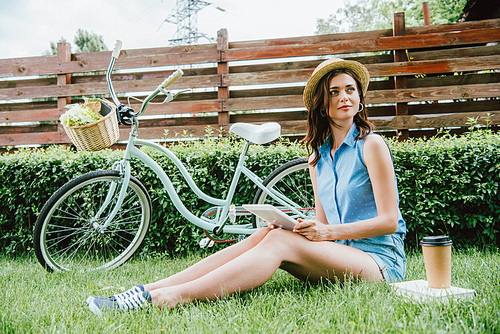 woman in straw hat holding digital tablet and sitting on grass near bicycle, paper cup and book