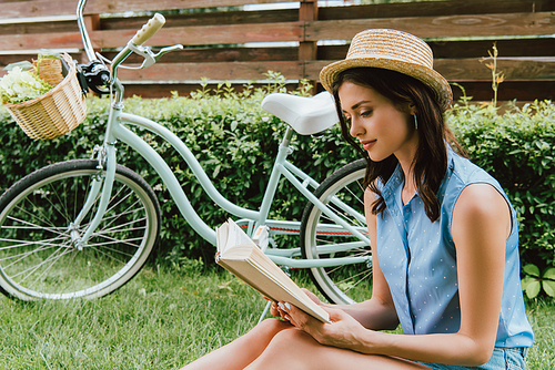 stylish girl in straw hat reading book and sitting on grass near bicycle