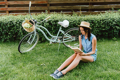 trendy girl in straw hat reading book and sitting on grass near bicycle with wicker basket