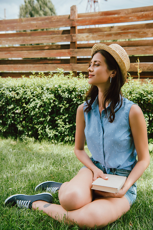 happy girl in straw hat holding book and sitting on grass