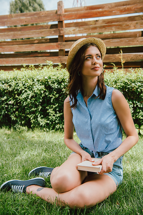 happy woman in straw hat holding book and sitting on green grass