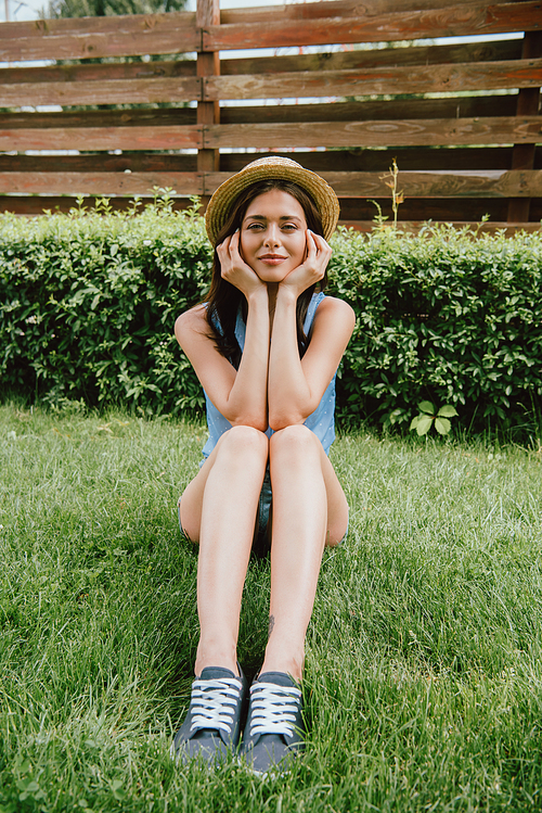 cheerful young woman in straw hat touching face and  while sitting on grass