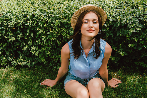 cheerful young woman in straw hat sitting on grass