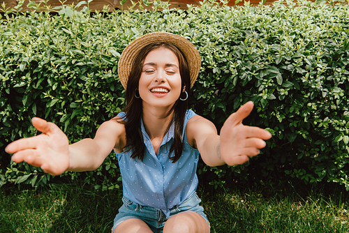 selective focus of happy girl in straw hat with outstretched hands sitting near bush