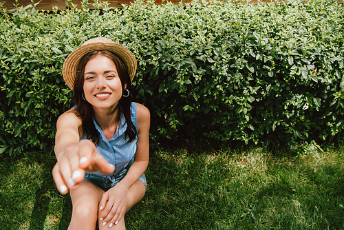 selective focus of happy girl in straw hat with outstretched hand sitting near bush