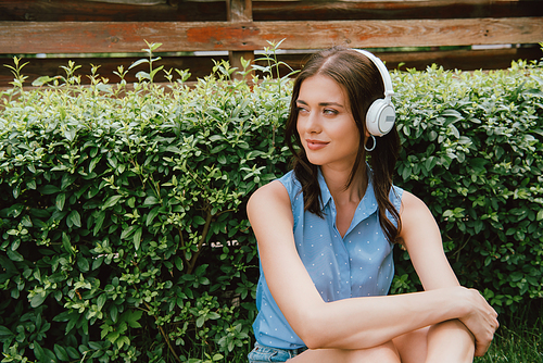 cheerful young woman listening music in wireless headphones near bush