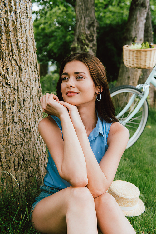 dreamy woman sitting on grass near bicycle and straw hat