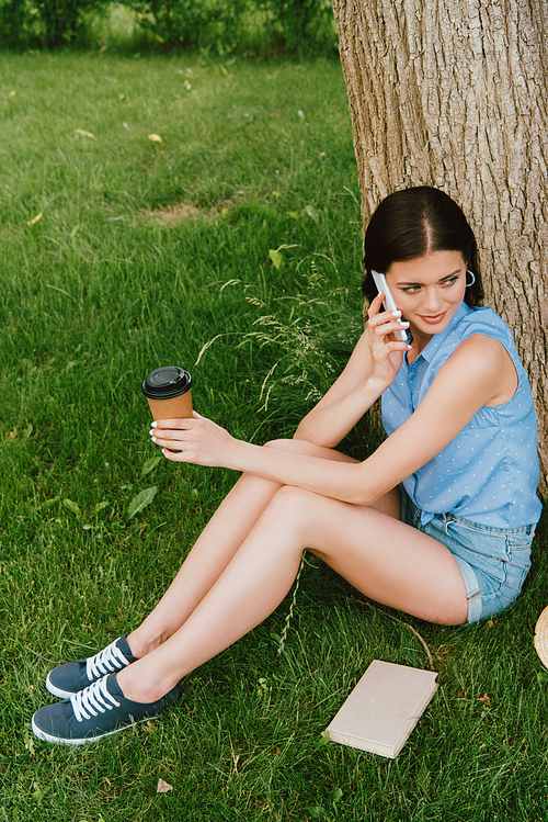 attractive woman talking on smartphone and holding paper cup while sitting on grass