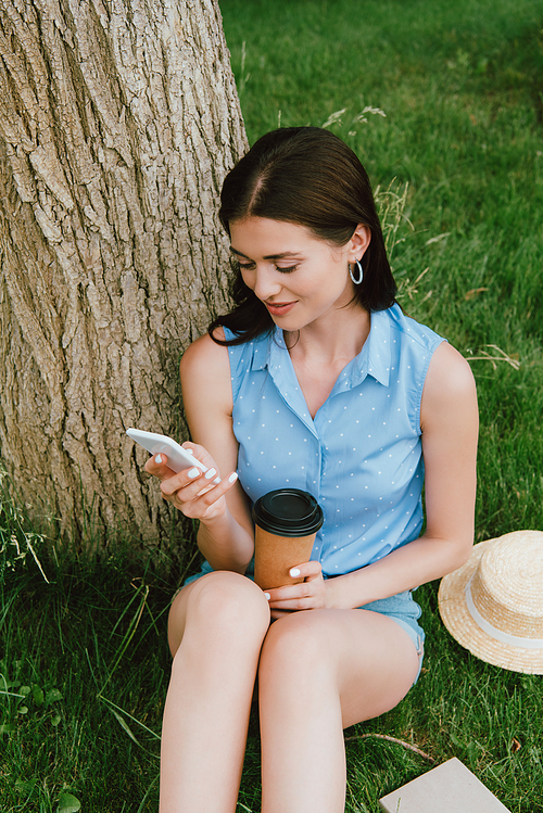 happy woman using smartphone and holding paper cup while sitting on grass