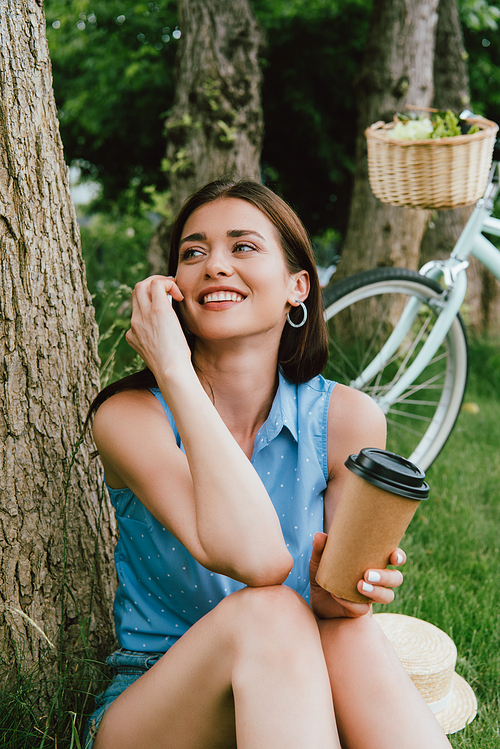 smiling woman talking on smartphone and holding paper cup while sitting near tree trunk