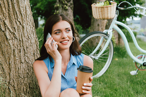 cheerful woman talking on smartphone and holding disposable cup outside