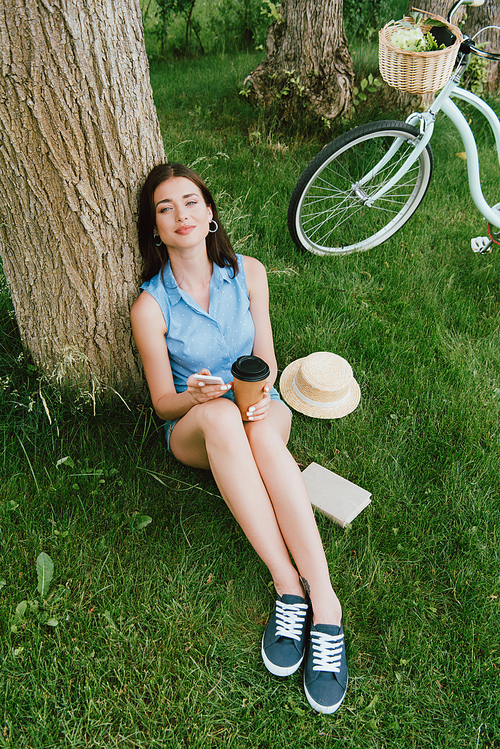 high angle view of beautiful woman smiling while holding smartphone and paper cup near tree trunk