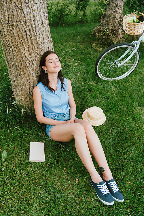 high angle view of happy woman sitting on grass near tree trunk, bicycle, book and straw hat