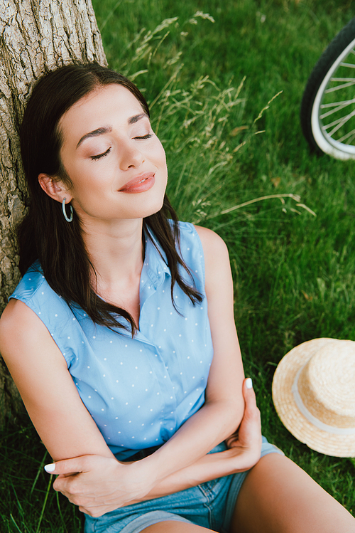 high angle view of beautiful woman smiling while sitting near tree trunk and straw hat