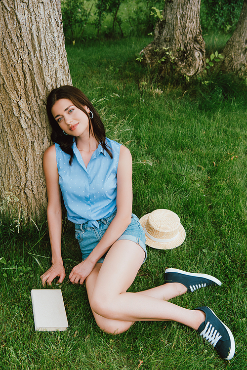 happy woman sitting on grass near tree trunks, book and straw hat