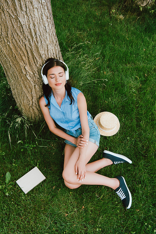 high angle view of young woman in closed eyes listening music while sitting on grass near book and stylish straw hat